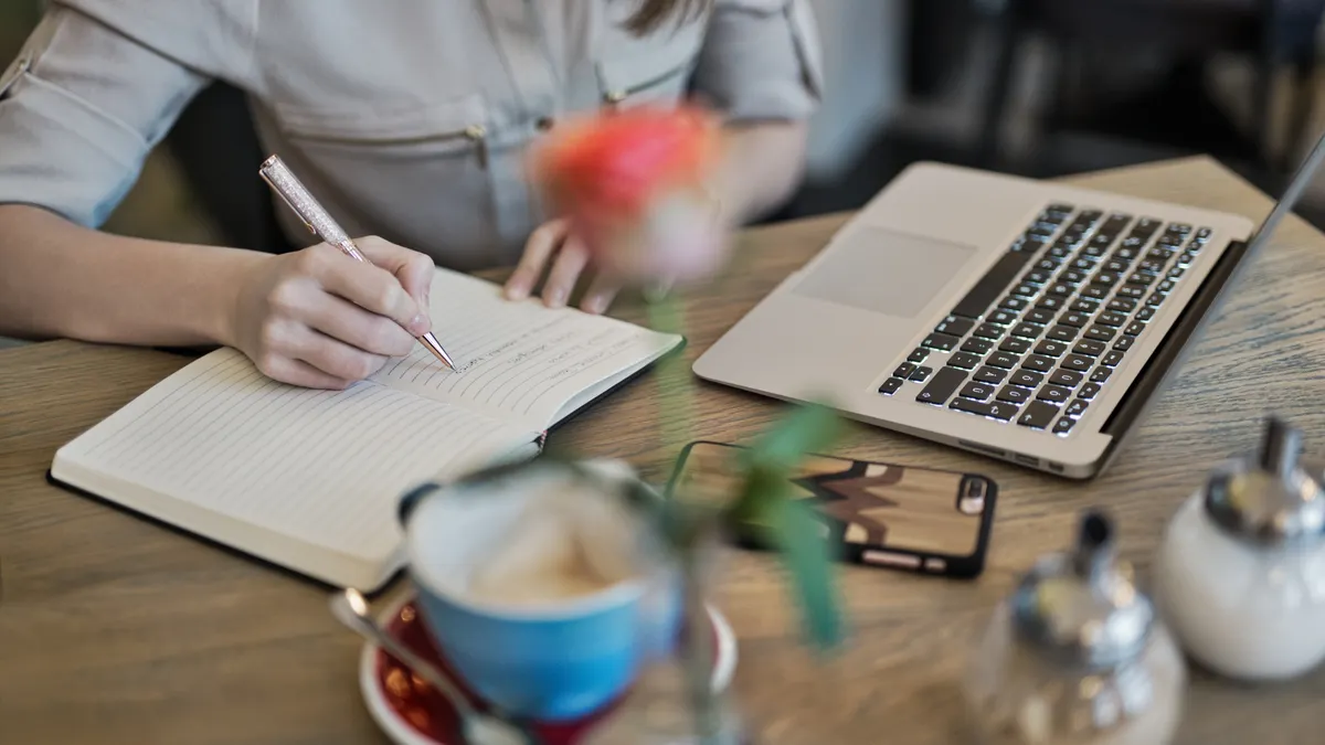 Person taking notes beside a laptop and phone