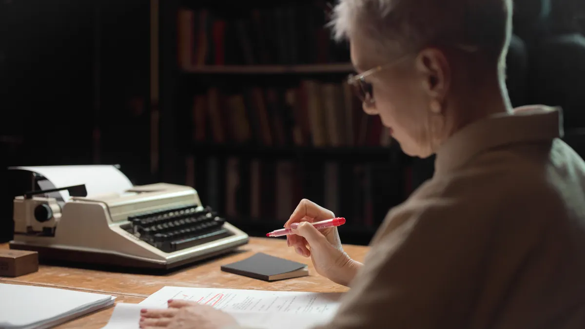 Older person writing beside a typewriter at a desk